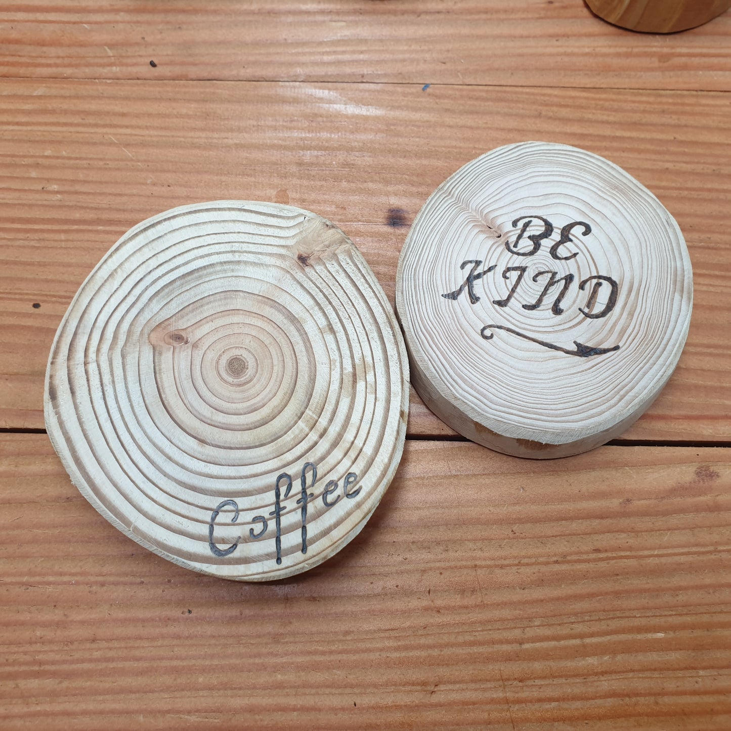 Two wooden coasters with 'Coffee' and 'Be Kind' text on a wooden surface.