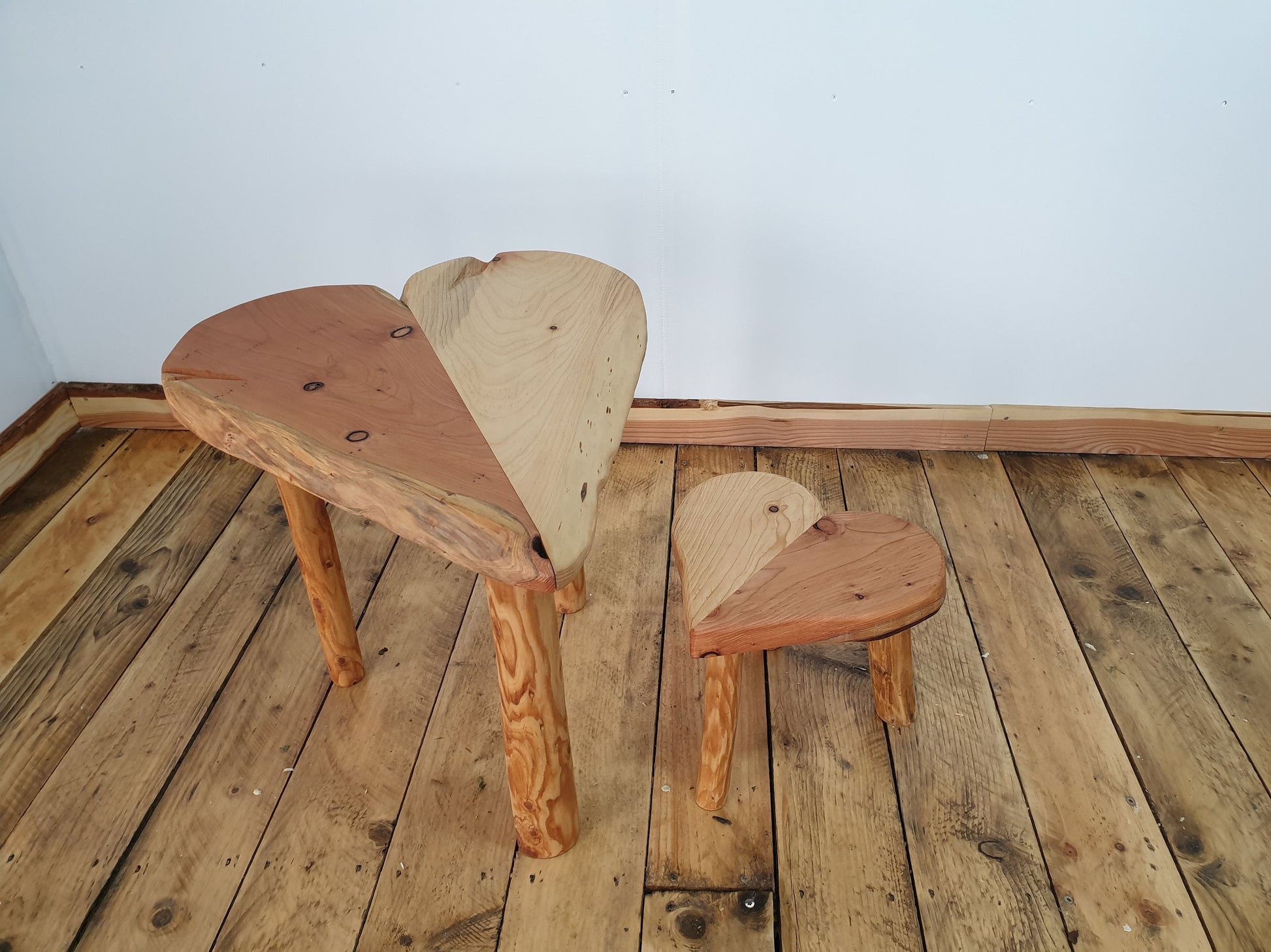 A photo of a heart shaped table and a small heart shaped stool. Half the hearts are made of red heart wood and the other halves are made from white wood giving it a striking unique look. Both supported by 3 peeled round legs.