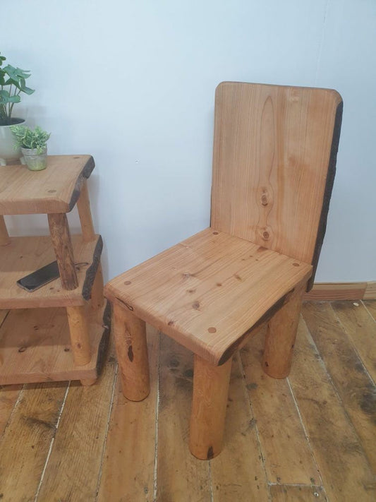 A short straight-backed wooden throne posed diagonally alongside a wooden end table with a potted plant atop it.