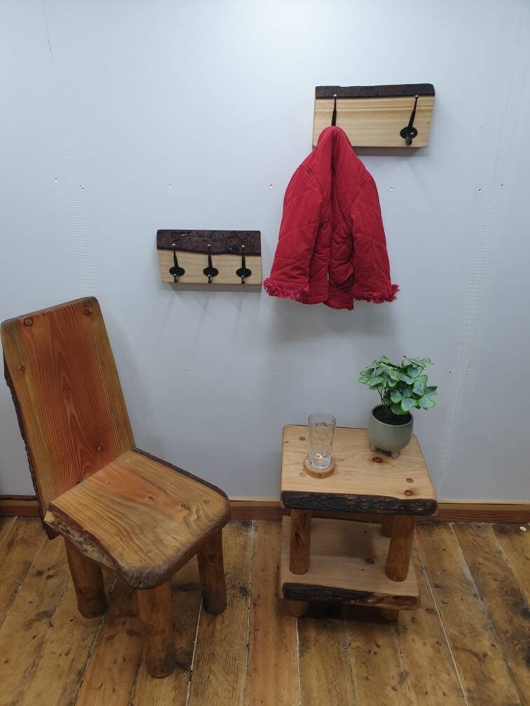 A photo of the same 2 previous coat racks mounted on a white wall but zoomed out to show a wooden chair and small wooden table that has a houseplant and a glass on a wooden coaster on it.