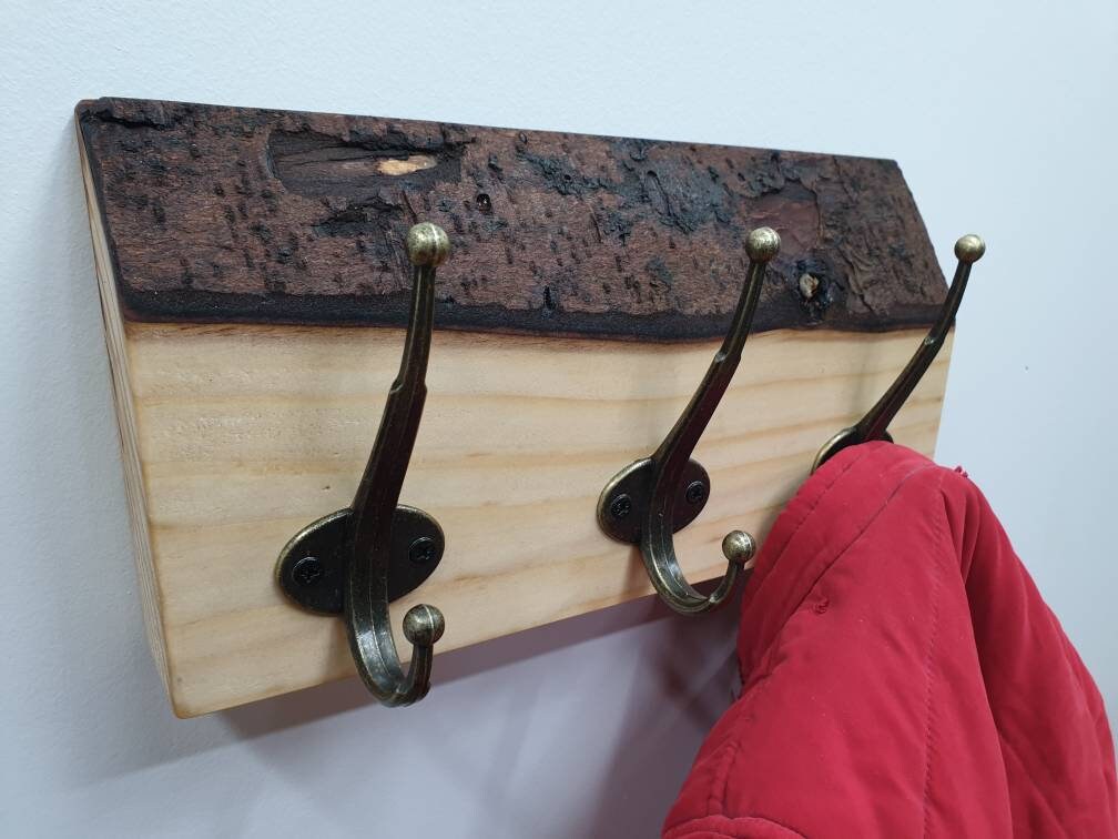 A close up photo of a 3 hook coat rack from the left aspect. It has a subtle creamy white wood grain and a chunky, dark chocolate colour bark layer along the top. a small red coat is hung of the right hand side hook.