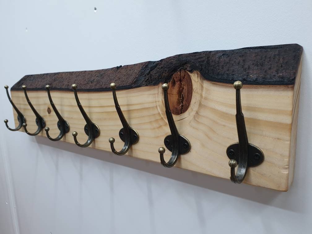 A 70 cm coat rack with 7 hooks from the left aspect mounted on a white wall. made from spruce it has a white sandy grain and bumpy, dark brown bark. It has a feature knot above the second from end hook that appears toffee brown in colour.