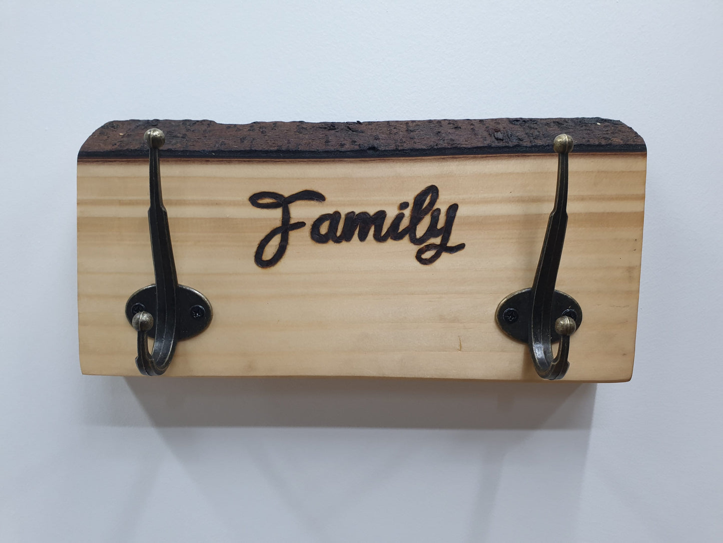 A small coat rack with 2 hooks with 'Family' engraved in between. The wood grain is very light in colour and its intact bark along the top edge is a dark chocolate brown.