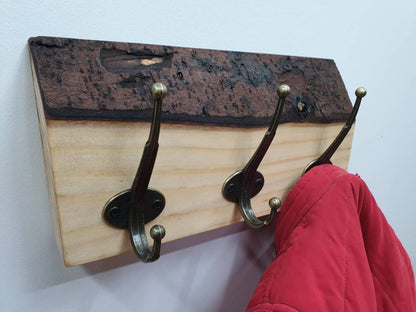 A close up photo of a 3 hook coat rack from the left aspect. It has a subtle creamy white wood grain and a chunky, dark chocolate colour bark layer along the top. a small red coat is hung of the right hand side hook.