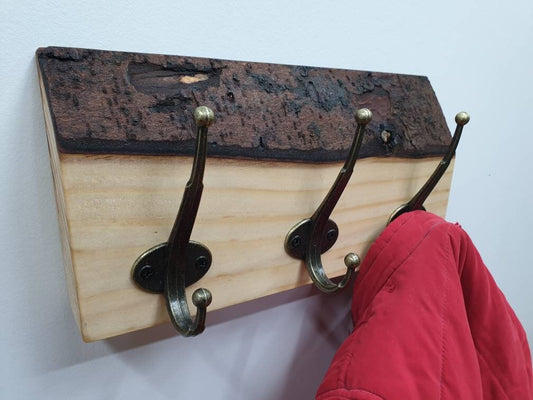 A close up photo of a 3 hook coat rack from the left aspect. It has a subtle creamy white wood grain and a chunky, dark chocolate colour bark layer along the top. a small red coat is hung of the right hand side hook.