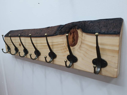 A 70 cm coat rack with 7 hooks from the left aspect mounted on a white wall. made from spruce it has a white sandy grain and bumpy, dark brown bark. It has a feature knot above the second from end hook that appears toffee brown in colour.