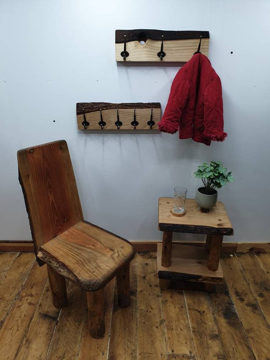 A photo of the same 2 previous coat racks mounted on a white wall, it is zoomed out to show a wooden chair and a small wooden table underneath. The table has a houseplant and a drinking glass on a wooden coaster. 