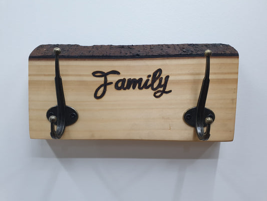 A small coat rack with 2 hooks with 'Family' engraved in between. The wood grain is very light in colour and its intact bark along the top edge is a dark chocolate brown.
